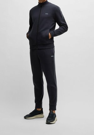 Mann trägt eine schwarze BOSS Markenzipjacke und passende Jogginghose mit Seitenstreifen sowie schwarze Sneaker, steht mit den Händen in den Taschen.