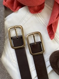 Kaksi tummanruskeaa nahkavyötä, joissa on suuret messinkisoljet, lepäävät valkoisella kangaspinnalla, vieressään punainen huivi ja muita tekstiilejä.