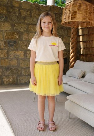 Junges Mädchen steht drinnen und trägt ein weißes T-Shirt mit Sonnengrafik und gelben Rock, pinke Sandalen, Steinmauer im Hintergrund, Korblampe in der Nähe.