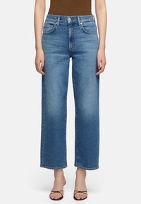 Femme portant un jean bleu taille haute à jambes droites et des sandales marron à lanières et bouts ouverts, debout devant un fond blanc uni.