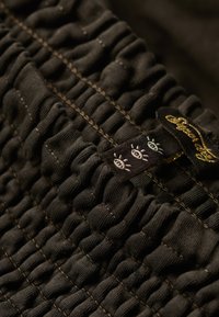 Tissu noir texturé avec des plis froncés et des coutures légères. L'étiquette visible présente un motif et des lettres dorées.