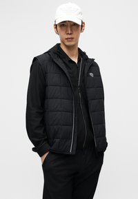 Μαύρο μπουφάν με επένδυση και κουκούλα, με φερμουάρ μπροστά και υφασμάτινες λεπτομέρειες. Συνδυασμένο με μαύρο πουκάμισο από δίχτυ και παντελόνι στο ίδιο χρώμα. Άσπρο καπέλο.