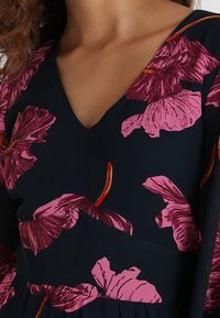 Bloemrijke jurk met een diepe V-hals. Bevat grote roze bloemenpatronen op een donkerblauwe achtergrond, met lange mouwen en een zachte, vloeiende textuur.