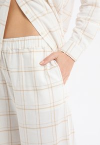 Komplet odzieży domowej w kratę w kolorze białym i beżowym, składający się z koszuli zapinanej na guziki oraz spodni z elastycznym pasem i bocznymi kieszeniami. Miękka, teksturowana tkanina.