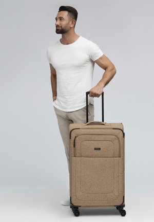 Valise en tissu marron avec un motif à carreaux, poignées rembourrées, poignée rétractable et quatre roues pour la mobilité. Le modèle se tient à côté.
