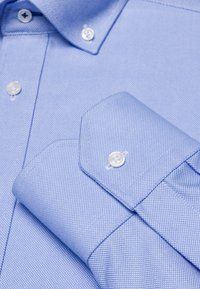 Camicia button-up di un blu chiaro con un motivo testurizzato, bottoni bianchi e un colletto a punta con polsini accentuati.