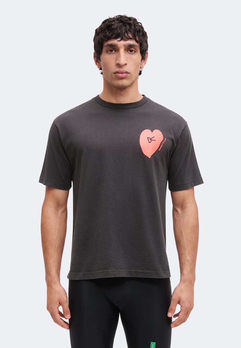 Άνδρας που φοράει σκούρο γκρι T-shirt με ένα μικρό ροζ καρδιά σχεδιασμό στο αριστερό στήθος, στέκεται μπροστά σε απλό φόντο.