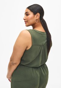 Jumpsuit senza maniche di un verde scuro con tessuto strutturato, design con schiena arricciata e vita elastica. Orecchini a cerchio completano l'aspetto.