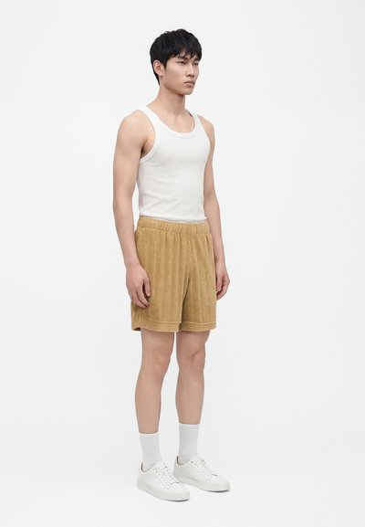 Jeune homme debout, portant un débardeur blanc, un short beige texturé, des chaussettes blanches et des baskets blanches, sur un fond clair uni.
