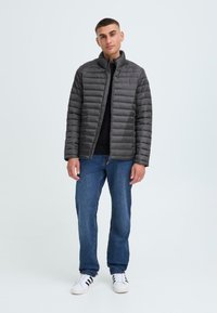 Grå pufferjakke med høy krage, med horisontalt quiltet design, båret over en svart genser, paret med blå jeans og hvite joggesko.