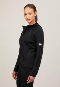 Giacca nera con cappuccio, zip frontale, tessuto texture e logo sulla manica. Presenta tasche laterali e una silhouette aderente.