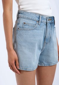 Shorts en denim bleu clair, avec une taille haute, cinq poches, un bouton en cuivre et des rivets, et une coupe décontractée avec un léger dégradé.