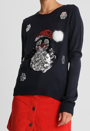 Pull bleu marine avec un pingouin en sequins portant un bonnet de Père Noël avec un pompon blanc, orné de flocons de neige argentés, porté avec une jupe rouge à boutons.