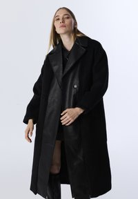 Schwarzer Mantel aus einer Kombination von Wolle und Leder, mit einem großen Kragen, doppelreihigen Knöpfen und einer strukturierten Oberfläche. Lange Ärmel und lockere Passform.