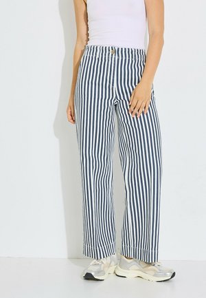 Mujer con pantalones de talle alto y pierna ancha con rayas verticales en azul marino y blanco, una camiseta blanca sin mangas y zapatillas beige.