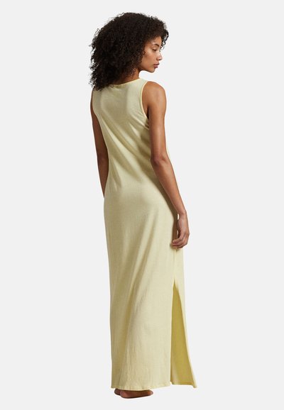 Femme aux cheveux bouclés portant une robe maxi jaune pâle sans manches avec une fente latérale, debout pieds nus, de profil avec la tête légèrement baissée.