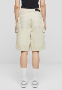 Beige cargo shortser med sidelommer og baklommer, laget av bomull, har en løs passform og oppbrettede buksebein. Brukt med hvite sneakers og sokker.