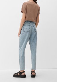 Hnědé tričko s krátkým rukávem ve spojení s vysokým pasem světlých modrých džínů a černými robustními sandály, které mají čistý design a mírně oprýskané lemy.