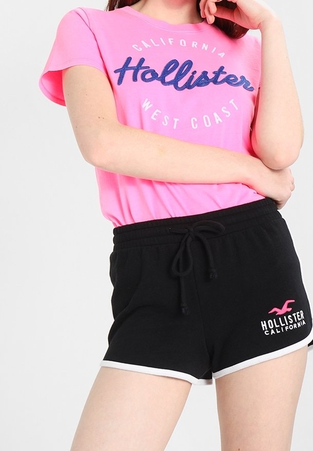 Růžové bavlněné tričko s modrým textem "Hollister West Coast", doplněné černými šortkami s bílým lemem a logem na levé noze.