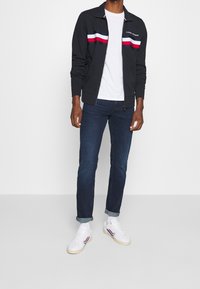 Černá zipová bunda s červenými, bílými a modrými pruhy, nošená přes bílou košili, v kombinaci s tmavě modrými džíny a bílými teniskami s pruhovanými akcenty.