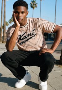 Nuori mies kyykyssä jalkakäytävällä, pukeutuneena beigeen kuviolliseen Kani-t-paitaan, mustiin housuihin ja valkoisiin tennareihin, taustalla palmupuita.