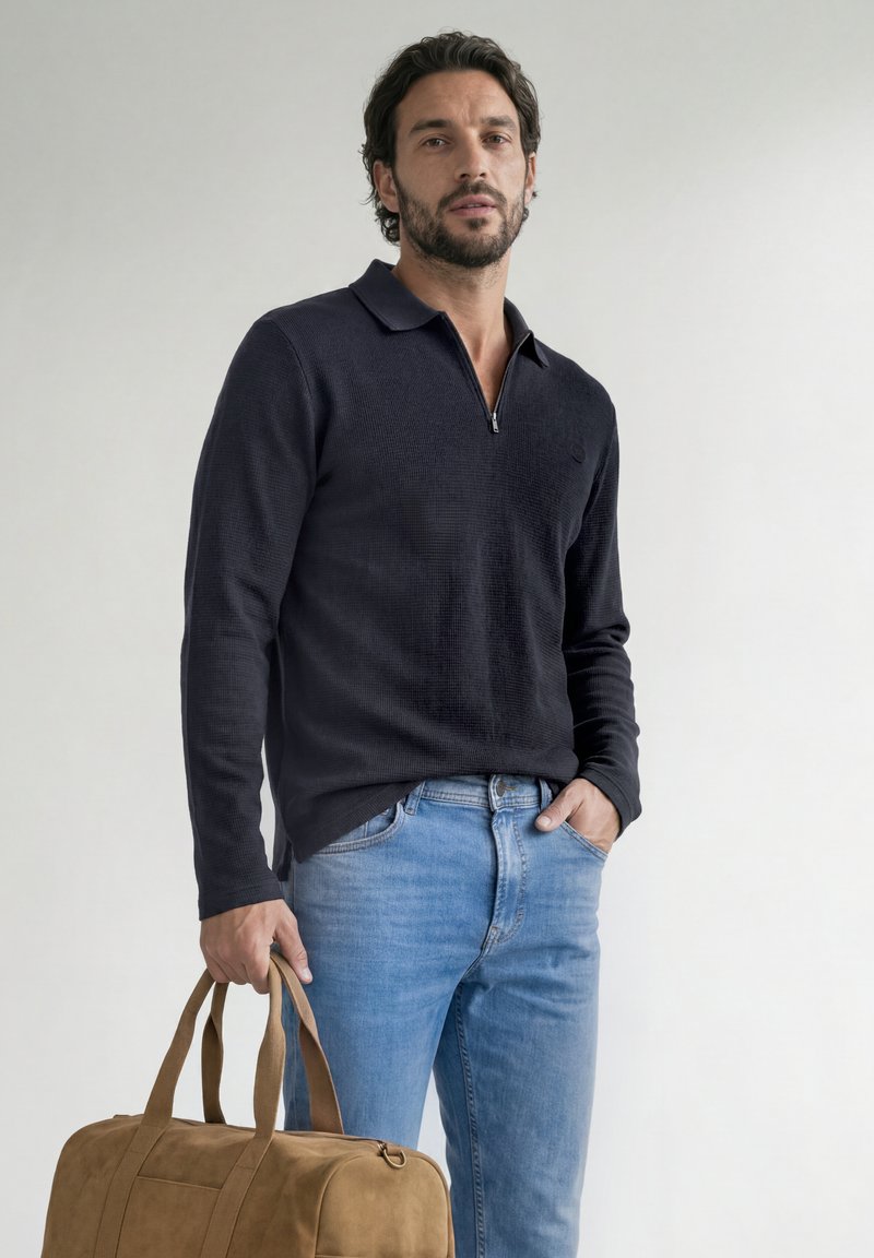 Homme aux cheveux courts et foncés avec une barbe, portant une chemise noire à manches longues et un jean bleu, tenant un sac de sport marron sur un fond clair.