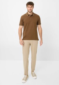 Polo shirt marrone con colletto e polsini a righe bianche, abbinato a pantaloni beige chiaro e sneakers beige, in piedi contro uno sfondo neutro.