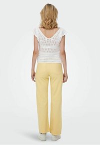 Biała koronkowa bluzka z krótkimi falbaaniastymi rękawami, zestawiona z żółtymi spodniami z wysokim stanem i białymi trampkami. Widok z tyłu ukazuje detale na ramionach i w talii.