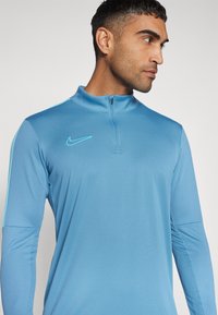 Nike hosszúujjú felső egyszínű világoskékben, magas gallérral, cipzáras elülső résszel és diszkrét logóval a bal mellen. Simább, rugalmas anyag.