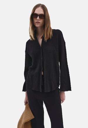 Zwarte shirt met textuur, met knoopsluiting aan de voorkant, lange mouwen en een losse pasvorm, gecombineerd met bijpassende broek en een gevlochten tan tas.