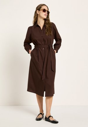 Frau trägt ein braunes Hemdkleid mit Gürtel und langen Ärmeln, Sonnenbrille und schwarze Mary Jane Schuhe, steht vor einem schlichten hellen Hintergrund.