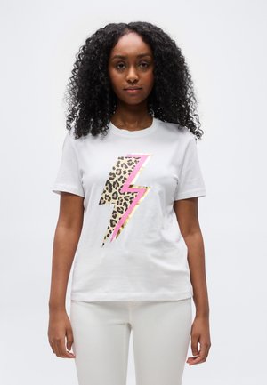 Kvinne med krøllete hår iført hvit t-skjorte med et leopardmønstret lyn i rosa og gull, kombinert med hvite bukser.