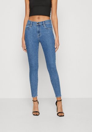 Vrouw draagt een blauwe slim fit jeans met hoge taille, een zwart cropped tanktop en zwarte hoge hakken met enkelbandjes, staande tegen een effen achtergrond.