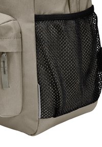 Sac à dos beige en toile texturée, avec une pochette latérale en filet noir et une fermeture éclair grise sur la poche avant.