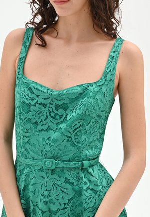 Femme portant une robe en dentelle verte sans manches avec un décolleté carré et une ceinture assortie sur un fond uni.