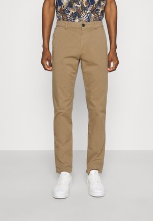 Mand iført slim-fit beige chinos, hvide sneakers og en kortærmet mønstret skjorte, står mod en helt hvid baggrund.