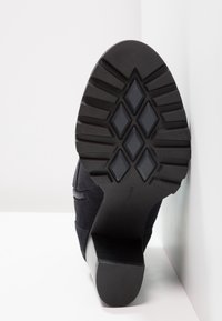 Černá semišová kotníková bota s texturovanou gumovou podrážkou, která má vrstvený podpatek a diagonální zipový detail po straně.