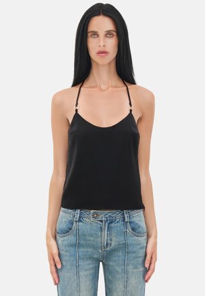 Donna con capelli lunghi e neri che indossa un top nero smanicato con scollo halter e jeans azzurri a vita alta su sfondo neutro.