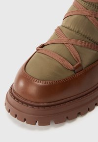 Καφέ δερμάτινη μ�πότα με επάνω μέρος από πράσινο ύφασμα, υφασμένη επιφάνεια και καφέ κορδόνια που διασταυρώνονται στην κορυφή. Επιθετικός σχεδιασμός λαστιχένιας σόλας.