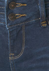 Vaqueros de mezclilla azul oscuro con costuras amarillas y dos botones metálicos visibles etiquetados "Productos de Mezclilla Genuine".