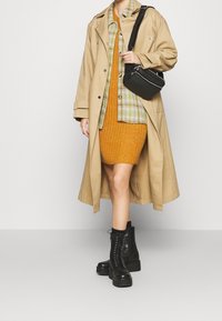 Manteau beige, chemise à carreaux verte et jaune, robe côtelée orange et bottines noires à lacets. Petit sac bandoulière noir inclus.