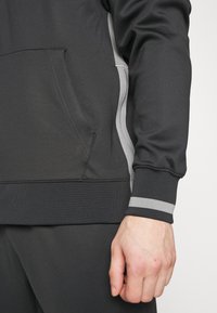 Sweatshirt de sport noir avec une fermeture éclair grise sur le côté et des rayures grises assorties au niveau des poignets. Comprend une poche avant et une finition texturée.