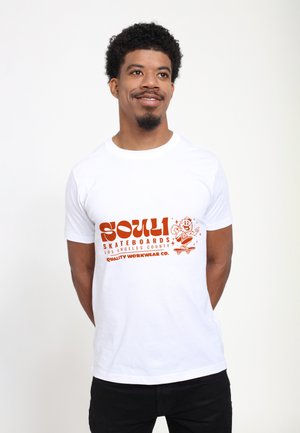 Ung mand med krøllet hår, der smiler, iført en hvid T-shirt med orange tekst "Souli Skateboards Los Angeles County" og en skateboarder-grafik.