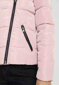 Veste courte en puffer rose clair avec texture matelassée, détails de fermeture éclair noirs et poches latérales. Tissu lisse avec une finition élégante.