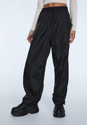 Femme portant un pantalon noir ample en nylon avec taille et poignets élastiques, des chaussures plateformes noires épaisses, et un haut blanc crop à manches longues.