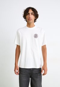 Hvid kortærmet t-shirt lavet af bomuld med rund halsudskæring. Har et cirkulært rødt og sort logo på venstre brystområde.