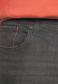 Tmavě šedé džínové kalhoty s klasickým střihem, vybavené přední kapsou a jemným prošíváním, v kombinaci s oranžovou mikinou.