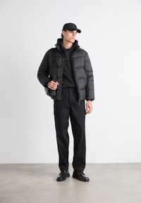 Svart pufferjakke med høy krage, lagdelt over en svart hettegenser. Sorte bukser og fottøy, med en svart caps og en messenger bag.