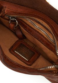 Interno di borsa in pelle marrone con fodera color cuoio, cerniera metallica e targhetta in pelle con dettagli in rilievo.