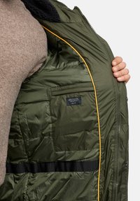 Zielona kurtka puchowa z teksturowanym wykończeniem, żółtymi lamówkami i kieszonką na piersi z etykietą z instrukcjami pielęgnacji, częściowo otwarta, aby pokazać szczegóły wnętrza.